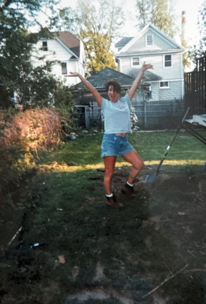 Scanned photo using iphone. Picture of a woman digging in the grass of a backyard in front of a weeping cherry tree.