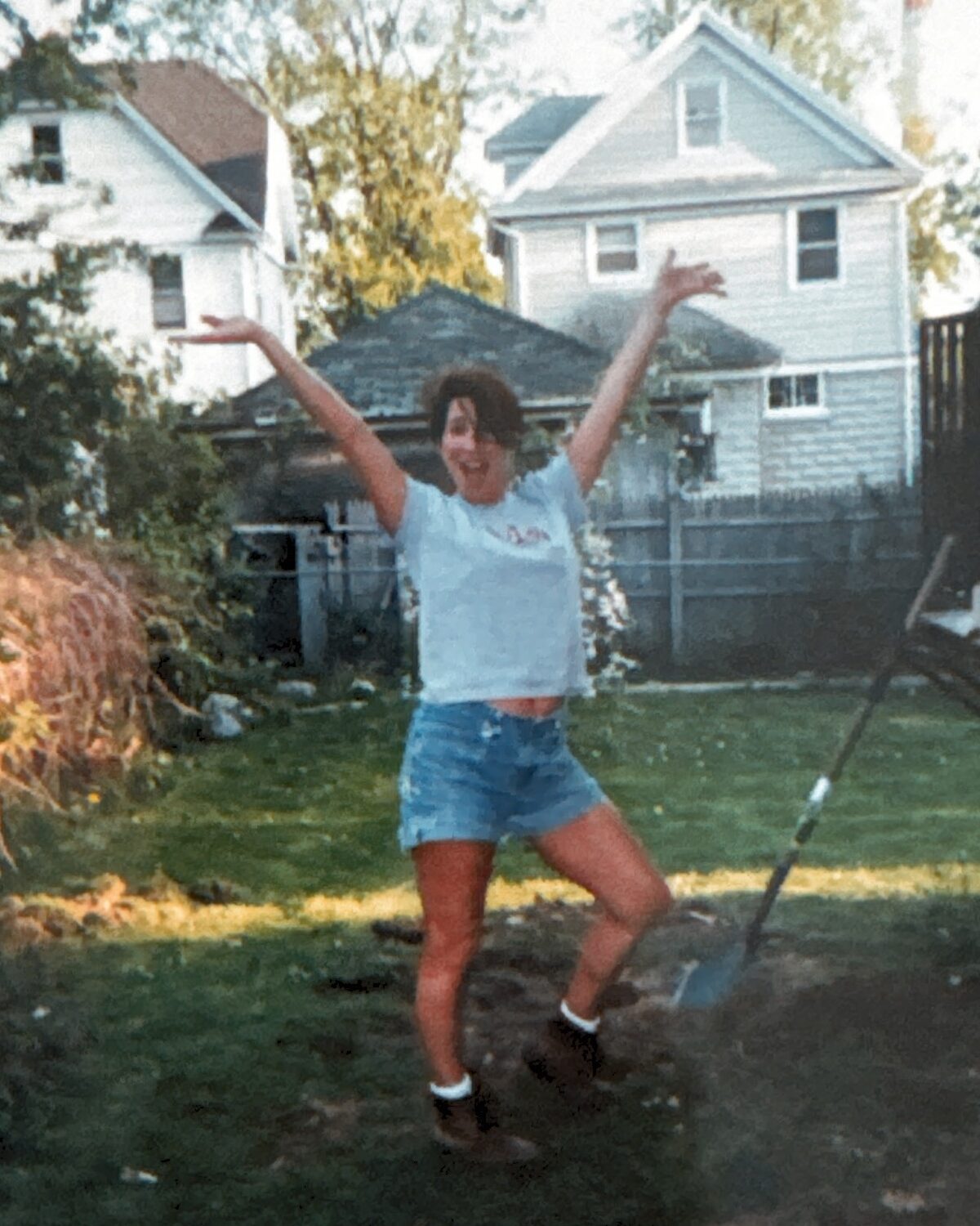 Scanned photo using iphone. Picture of a woman digging in the grass of a backyard in front of a weeping cherry tree.