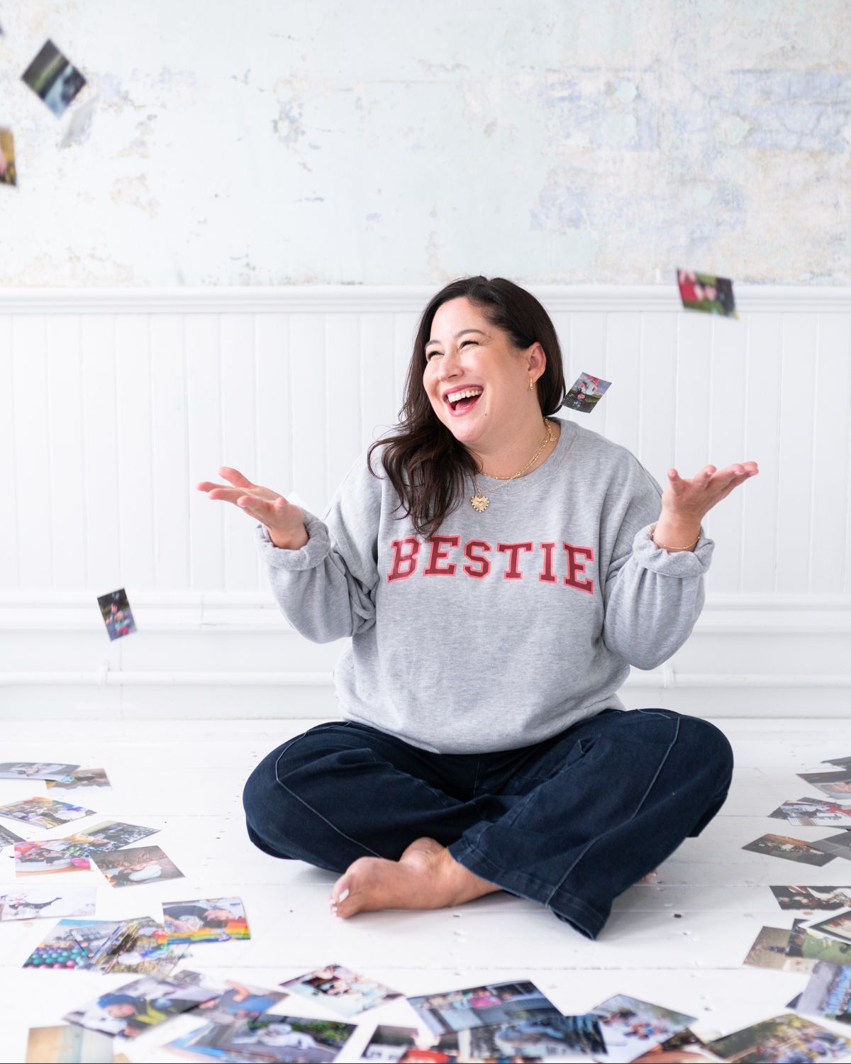 photo organizer Kiera Liu sitting on the floor in a pile of photographs. She is smiling and laughing and is wearing a Bestie sweatshirt.