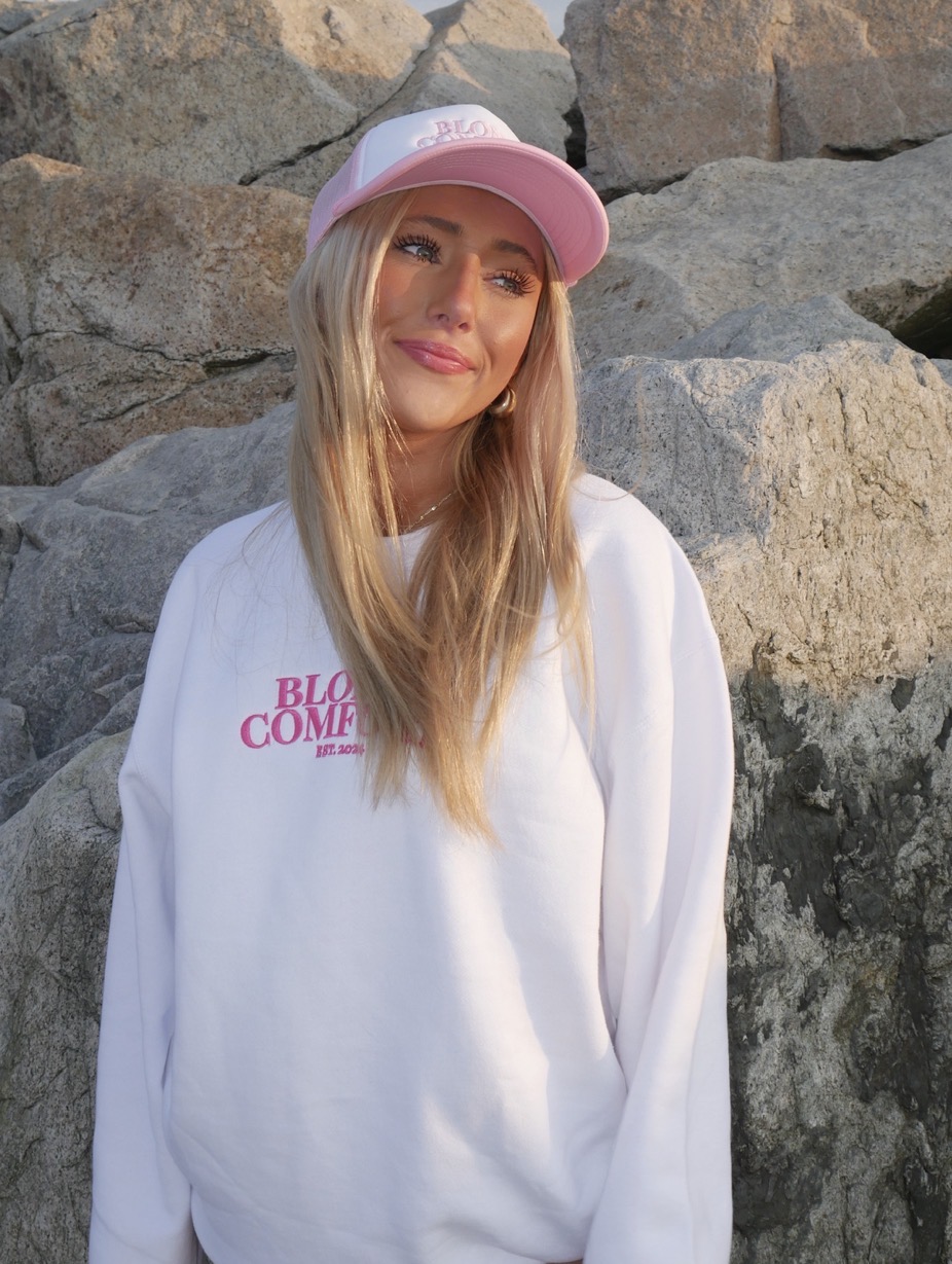 Blondi founder, Zoe Garcia wearing a pink hat and white sweatshirt she designed. Standing in front of a rock wall